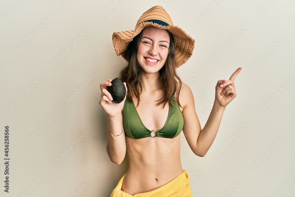 Young brunette woman holding avocado smiling happy pointing with hand and finger to the side Young brunette woman holding avocado smiling happy pointing with hand and finger to the side