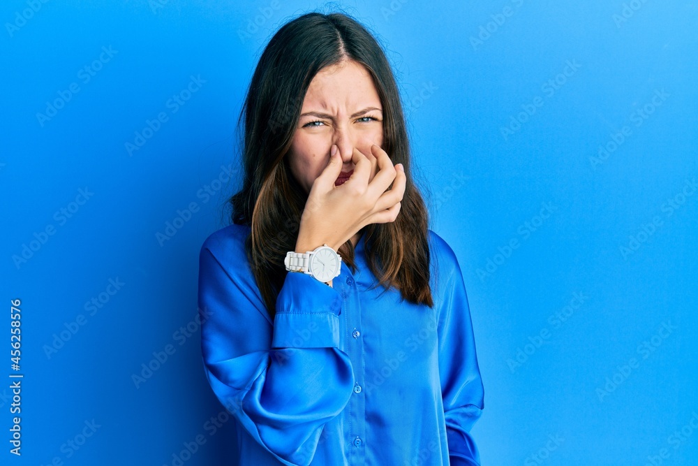 Young brunette woman wearing casual blue shirt smelling something stinky and disgusting, intolerable smell, holding breath with fingers on nose. bad smell Young brunette woman wearing casual blue shirt smelling something stinky and disgusting, intolerable smell, holding breath with fingers on nose. bad smell