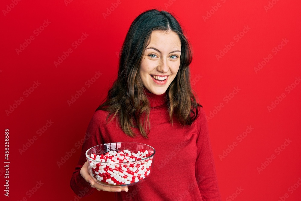 Young brunette woman holding bowl full of pills looking positive and happy standing and smiling with a confident smile showing teeth Young brunette woman holding bowl full of pills looking positive and happy standing and smiling with a confident smile showing teeth