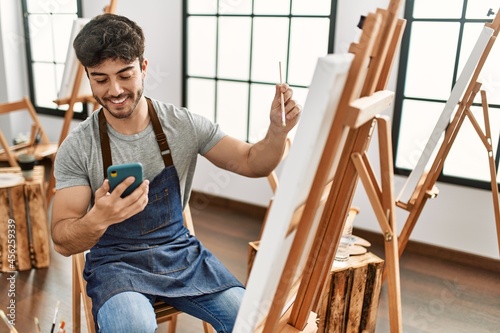 Young hispanic artist man smiling happy painting and using smartphone at art studio.
