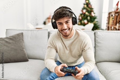 Young hispanic man smiling happy sitting on the sofa playing video game at home.