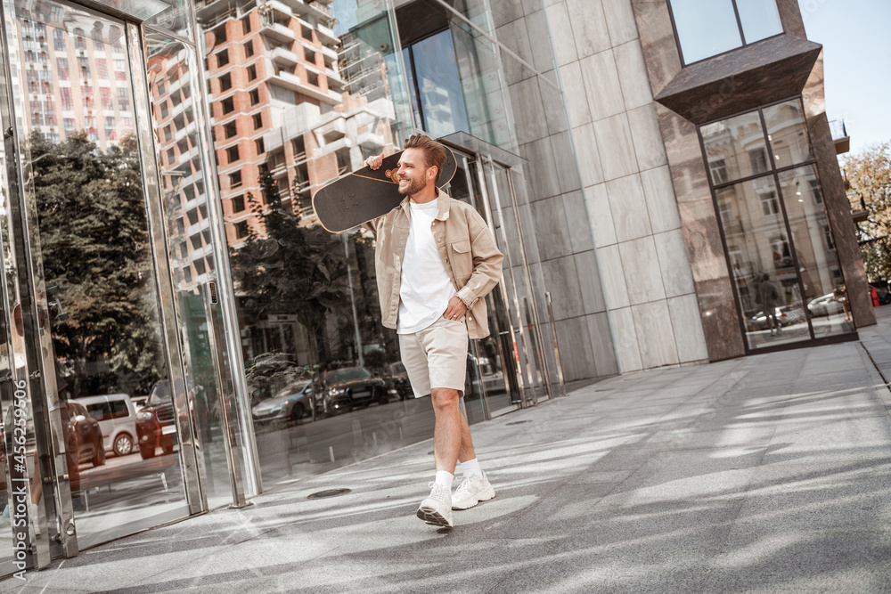 Attractive smiling young blonde man walks holding skateboard on shoulder at city street among glass buildings. Wears denim outfit. Sporty stylish skateboarder thinking about extreme trick outside.