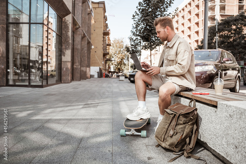 Young blonde man skater sitting on bench with laptop smartphone communicating online, streaming with longboard and backpack takeaway coffee. Remote work on the go. Busy city life. Stylish millennial.