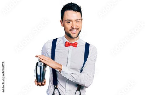 Hispanic man with beard preparing cocktail mixing drink with shaker winking looking at the camera with sexy expression, cheerful and happy face.
