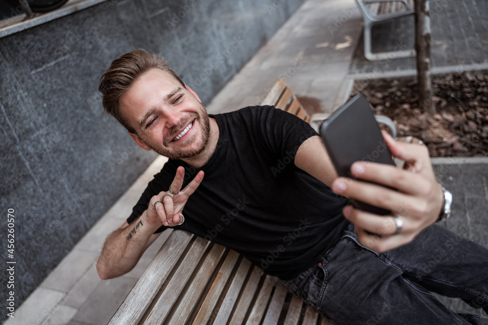 Handsome laughing young blonde man making selfie or streaming live on smartphone and gesturing peace sign while sitting on the bench on the street wears black jeans and black t-shirt