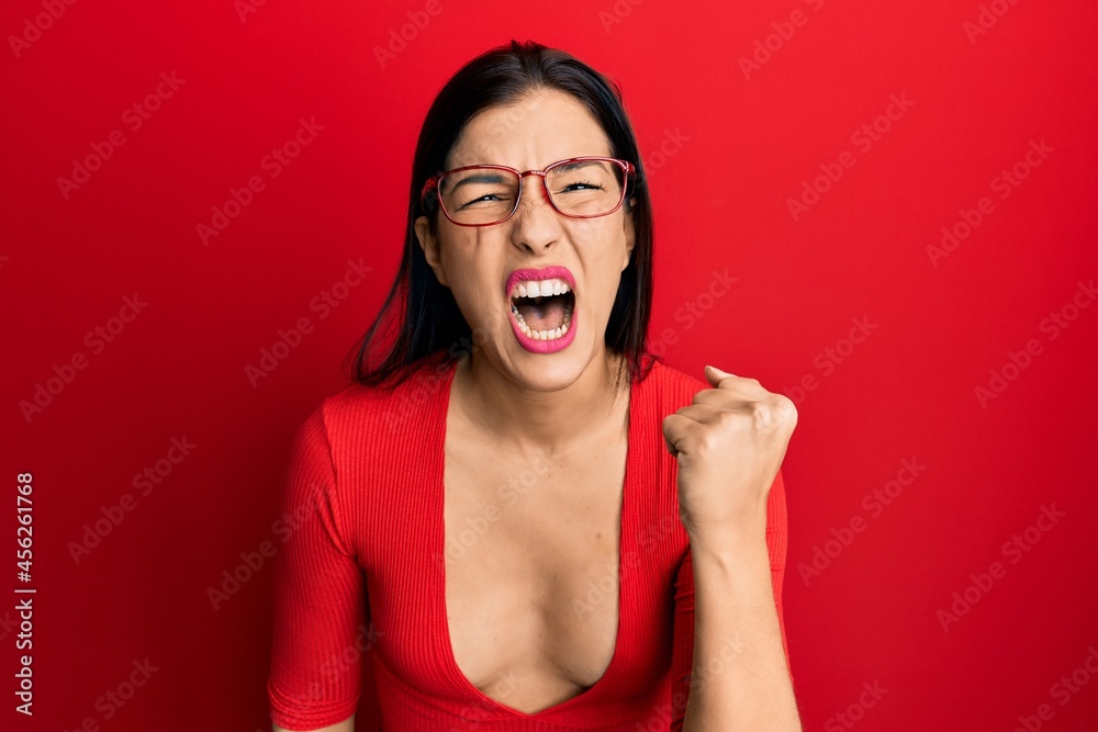 Young latin woman wearing casual clothes and glasses angry and mad raising fist frustrated and furious while shouting with anger. rage and aggressive concept. Young latin woman wearing casual clothes and glasses angry and mad raising fist frustrated and furious while shouting with anger. rage and aggressive concept.