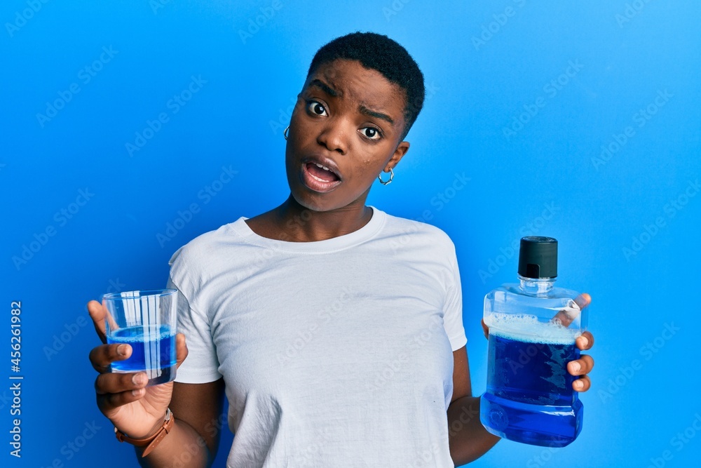 Young african american woman holding mouthwash for fresh breath in shock face, looking skeptical and sarcastic, surprised with open mouth Young african american woman holding mouthwash for fresh breath in shock face, looking skeptical and sarcastic, surprised with open mouth