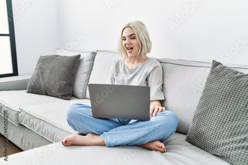 Young caucasian woman using laptop at home sitting on the sofa winking looking at the camera with sexy expression, cheerful and happy face.