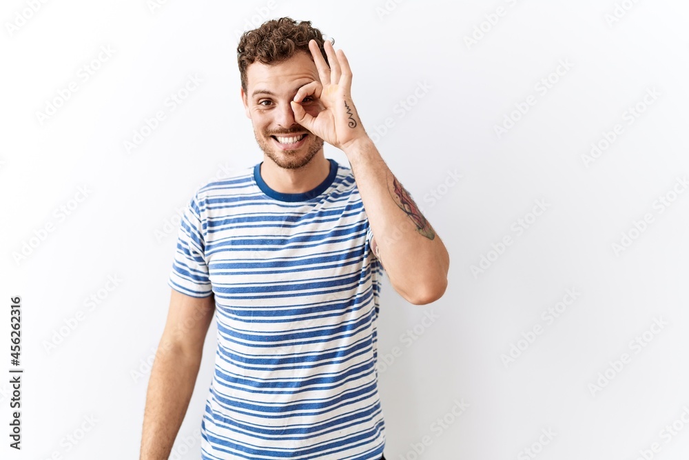 Handsome young man standing over isolated background doing ok gesture with hand smiling, eye looking through fingers with happy face. Handsome young man standing over isolated background doing ok gesture with hand smiling, eye looking through fingers with happy face.