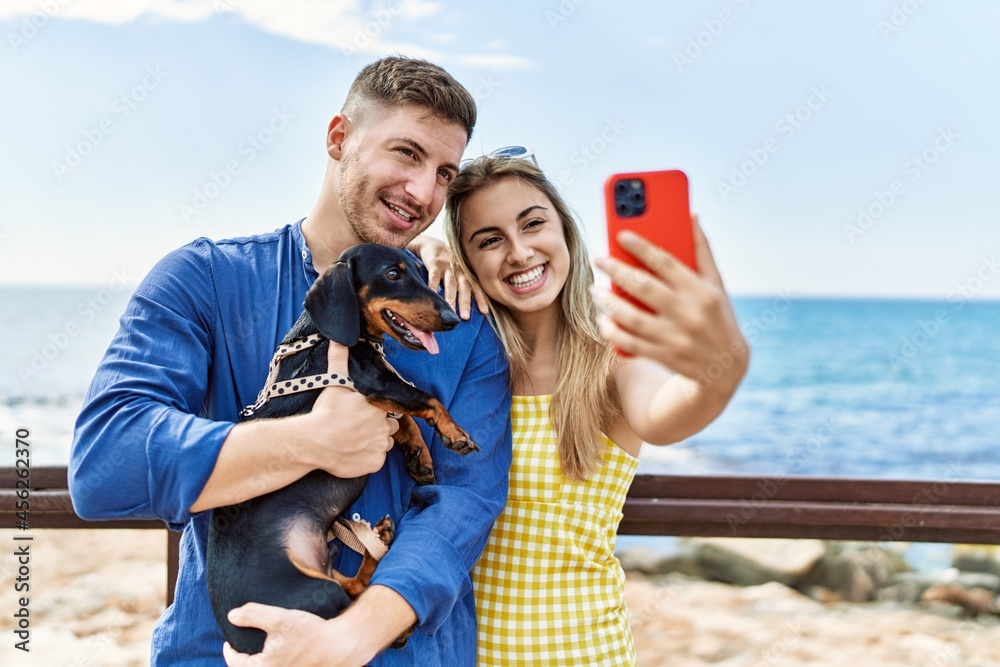 Young caucasian couple standing with dog make selfie by the smartphone at the beach.