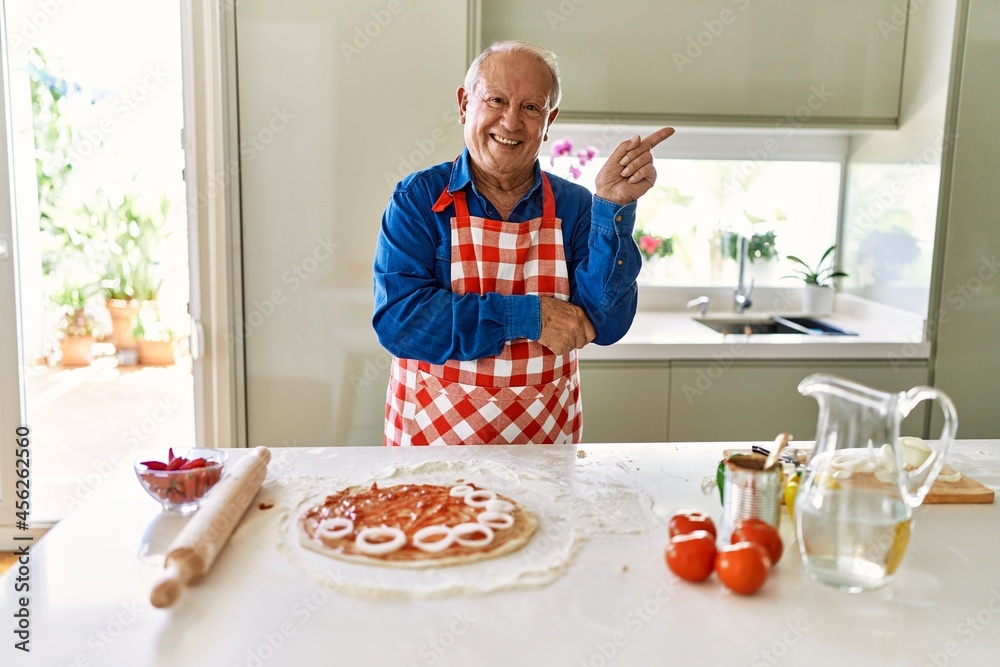 Senior man with grey hair cooking pizza at home kitchen smiling happy pointing with hand and finger to the side Senior man with grey hair cooking pizza at home kitchen smiling happy pointing with hand and finger to the side