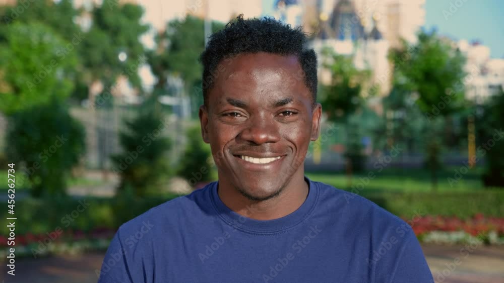 Close up smiling afro-american man looks camera on background beautiful park in summer. Portrait happy African male student wears blue t-shirt Close up smiling afro-american man looks camera on background beautiful park in summer. Portrait happy African male student wears blue t-shirt