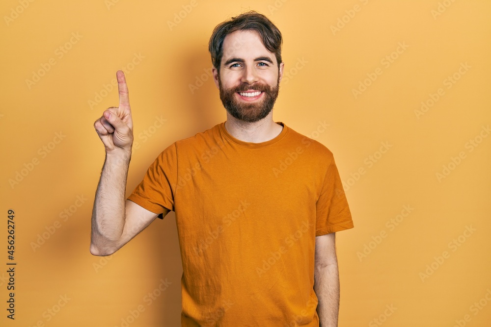 Caucasian man with beard wearing casual yellow t shirt showing and pointing up with finger number one while smiling confident and happy. Caucasian man with beard wearing casual yellow t shirt showing and pointing up with finger number one while smiling confident and happy.