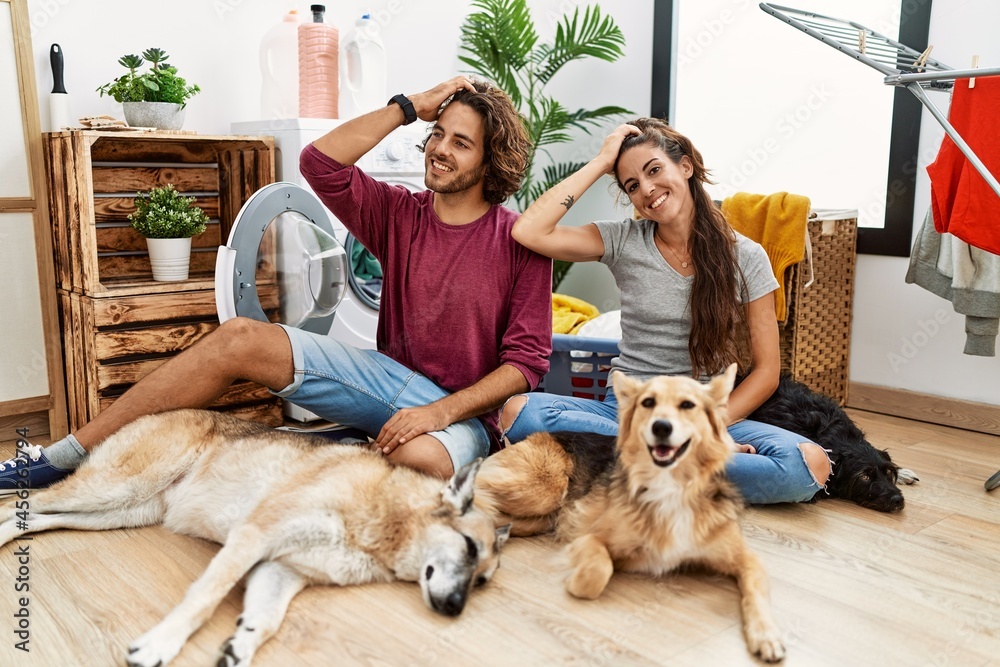 Young hispanic couple doing laundry with dogs smiling confident touching hair with hand up gesture, posing attractive and fashionable