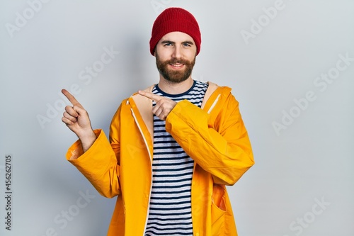 Caucasian man with beard wearing yellow raincoat smiling and looking at the camera pointing with two hands and fingers to the side.