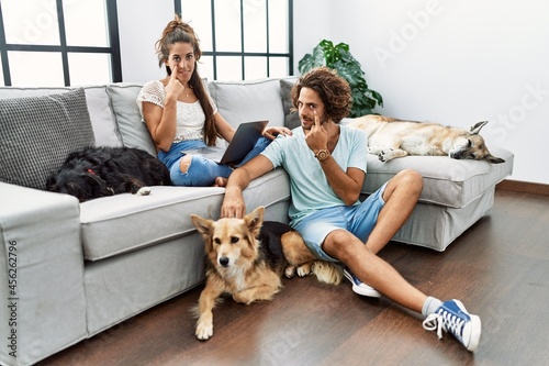 Young hispanic couple with dogs relaxing at home pointing to the eye watching you gesture, suspicious expression