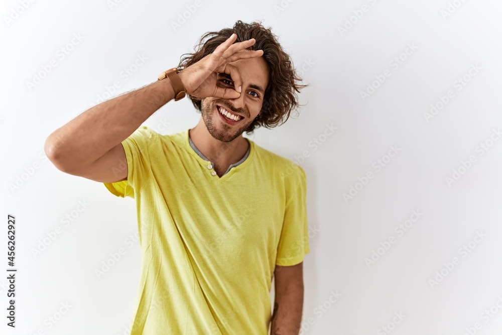 Young hispanic man standing over isolated background doing ok gesture with hand smiling, eye looking through fingers with happy face. Young hispanic man standing over isolated background doing ok gesture with hand smiling, eye looking through fingers with happy face.