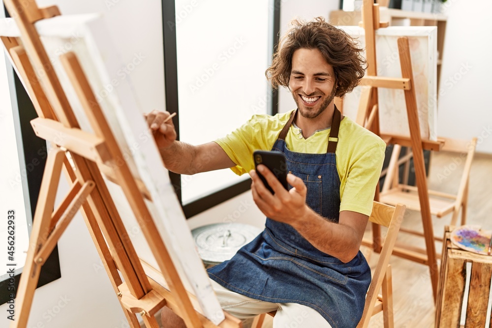 Young hispanic artist man smiling happy drawing and using smartphone at art studio.