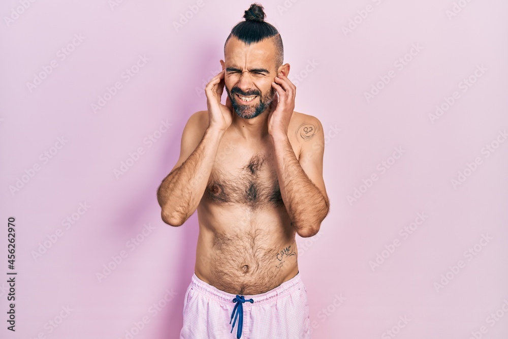 Fototapeta premium Young hispanic man shirtless wearing swimsuit covering ears with fingers with annoyed expression for the noise of loud music. deaf concept.