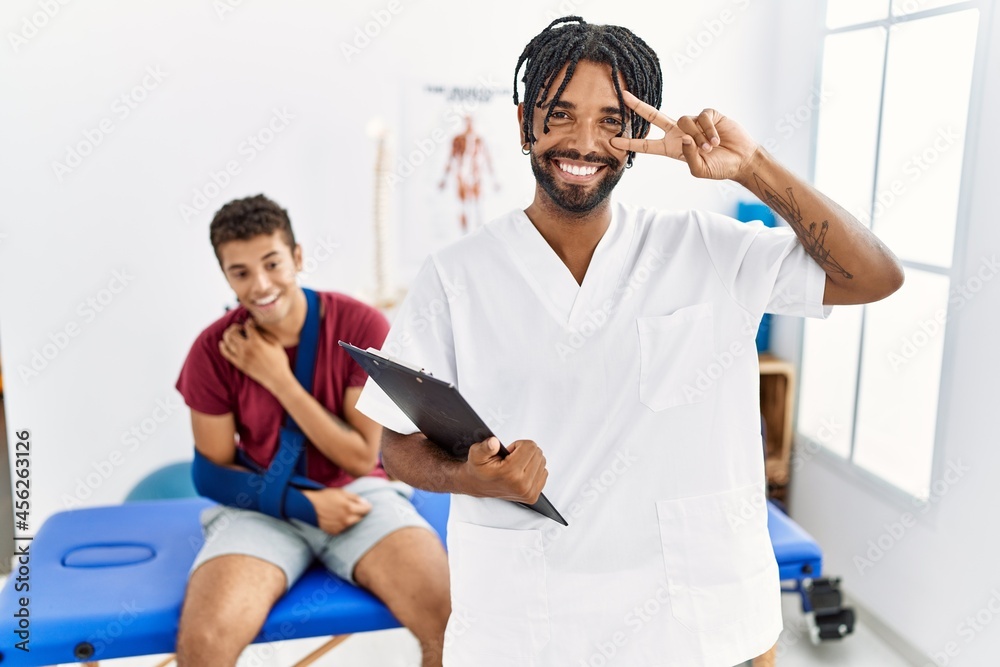 Obraz premium Young hispanic man working at pain recovery clinic with a man with broken arm doing peace symbol with fingers over face, smiling cheerful showing victory