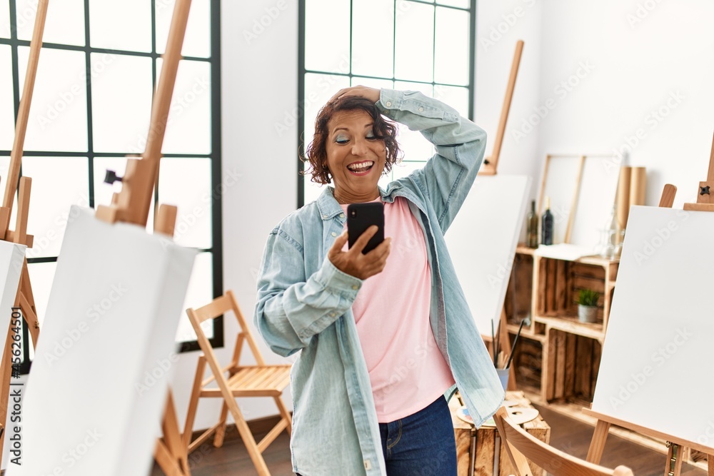 Middle age hispanic artist woman smiling happy having video call using smartphone at art studio.