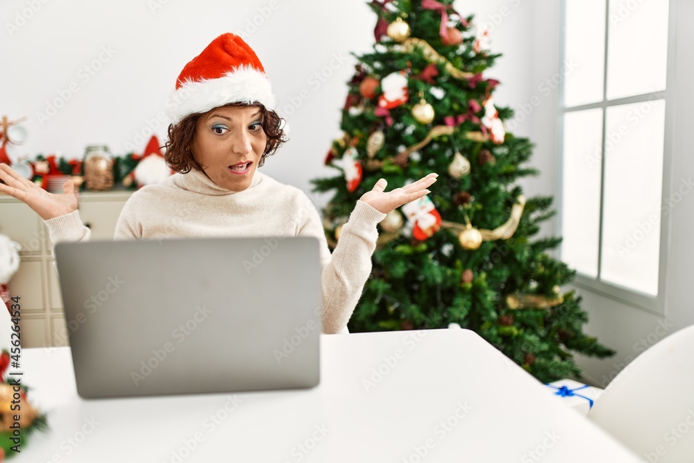 Middle age hispanic woman using laptop sitting by christmas tree clueless and confused expression with arms and hands raised. doubt concept. Middle age hispanic woman using laptop sitting by christmas tree clueless and confused expression with arms and hands raised. doubt concept.