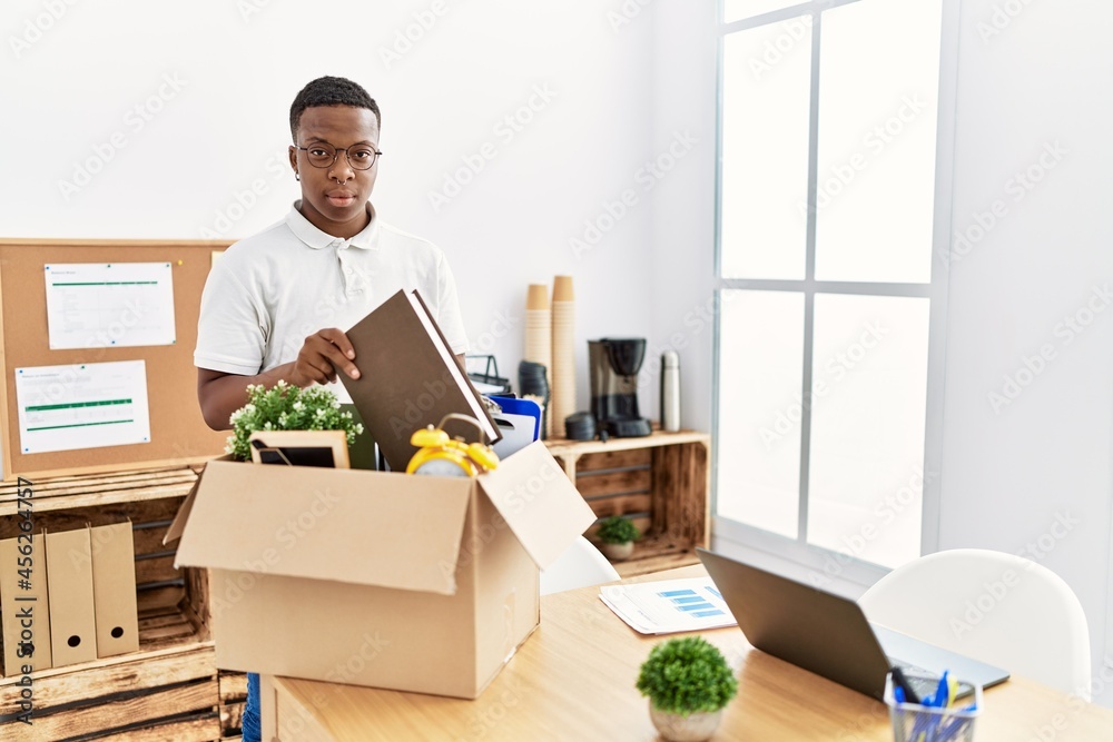 Young african man putting office objects into cardboard box thinking attitude and sober expression looking self confident Young african man putting office objects into cardboard box thinking attitude and sober expression looking self confident