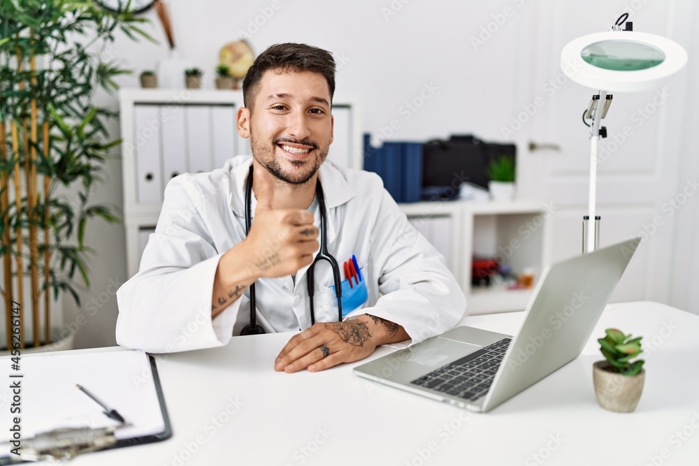 Young doctor working at the clinic using computer laptop doing happy thumbs up gesture with hand. approving expression looking at the camera showing success. Young doctor working at the clinic using computer laptop doing happy thumbs up gesture with hand. approving expression looking at the camera showing success.
