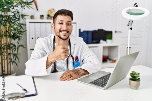 Young doctor working at the clinic using computer laptop doing happy thumbs up gesture with hand. approving expression looking at the camera showing success.
