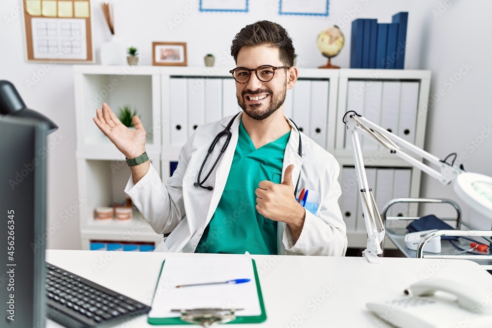 Young man with beard wearing doctor uniform and stethoscope at the clinic showing palm hand and doing ok gesture with thumbs up, smiling happy and cheerful Young man with beard wearing doctor uniform and stethoscope at the clinic showing palm hand and doing ok gesture with thumbs up, smiling happy and cheerful
