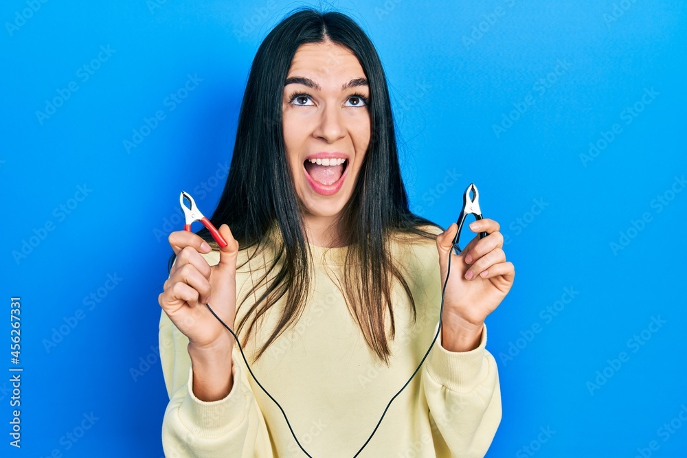 Obraz premium Young brunette woman holding battery clamps angry and mad screaming frustrated and furious, shouting with anger looking up.