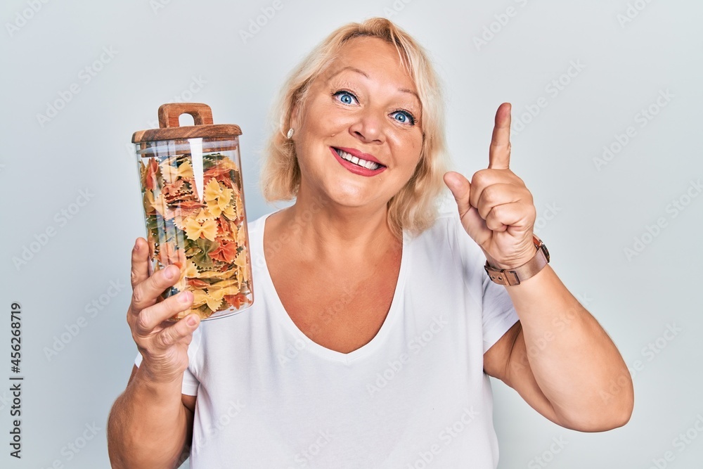 Middle age blonde woman holding jar with uncooked pasta smiling with an idea or question pointing finger with happy face, number one Middle age blonde woman holding jar with uncooked pasta smiling with an idea or question pointing finger with happy face, number one