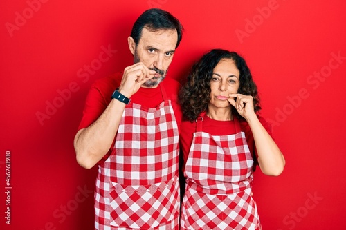 Middle age couple of hispanic woman and man wearing cook apron mouth and lips shut as zip with fingers. secret and silent, taboo talking