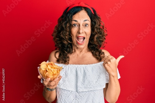 Middle age hispanic woman holding potato chip pointing thumb up to the side smiling happy with open mouth