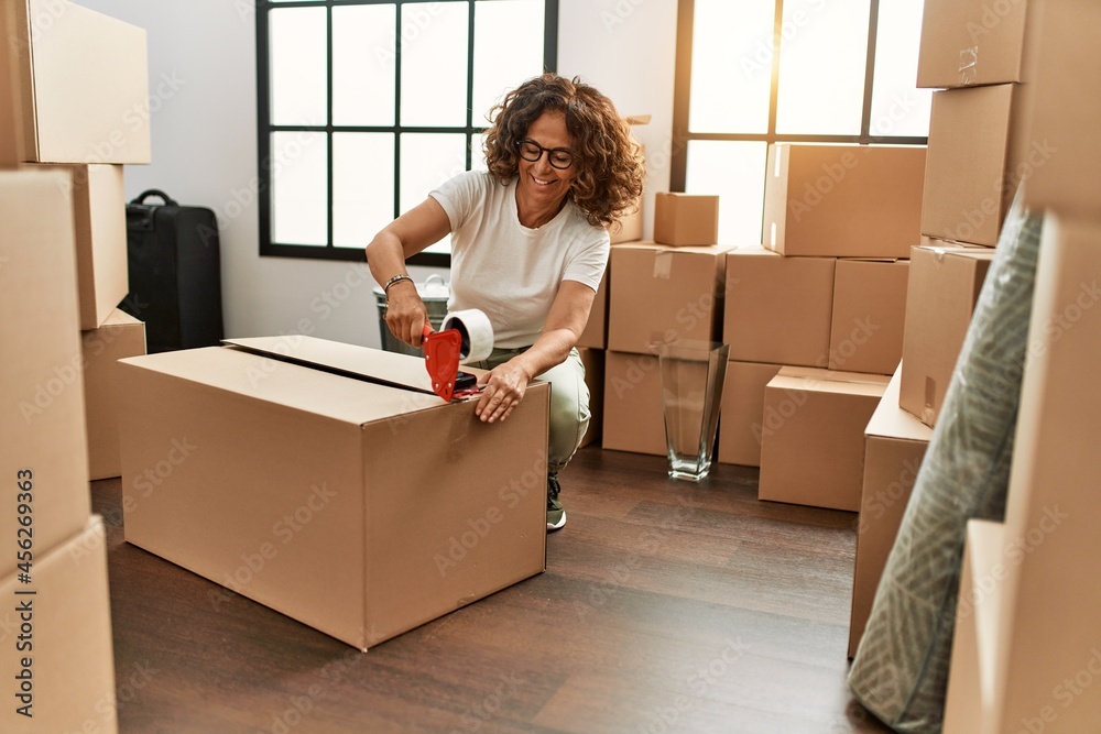 Fototapeta premium Middle age hispanic woman smiling confident packing cardboard box at new home