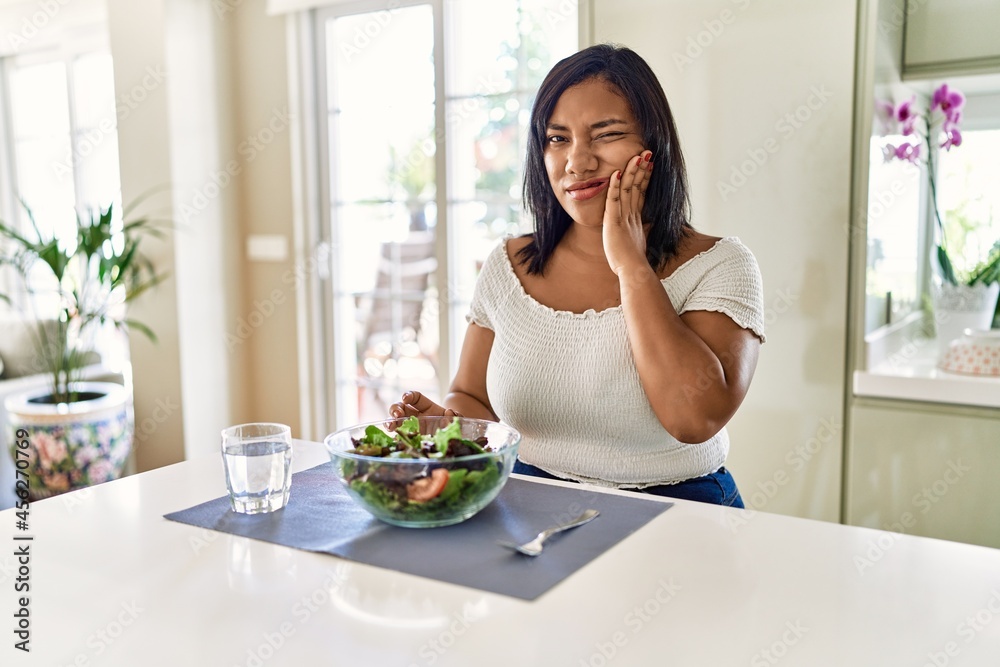 Young hispanic woman eating healthy salad at home touching mouth with hand with painful expression because of toothache or dental illness on teeth. dentist