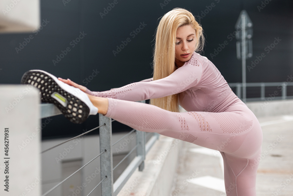 Beautiful sporty blonde girl with slim body in fashion pink sportswear doing leg stretching and warm up in the city Beautiful sporty blonde girl with slim body in fashion pink sportswear doing leg stretching and warm up in the city