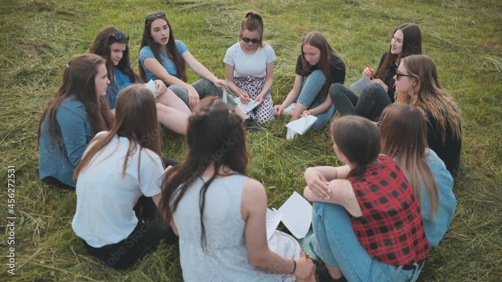 A group of female students are sitting in a circle on a meadow for collective work with notebooks. A group of female students are sitting in a circle on a meadow for collective work with notebooks.