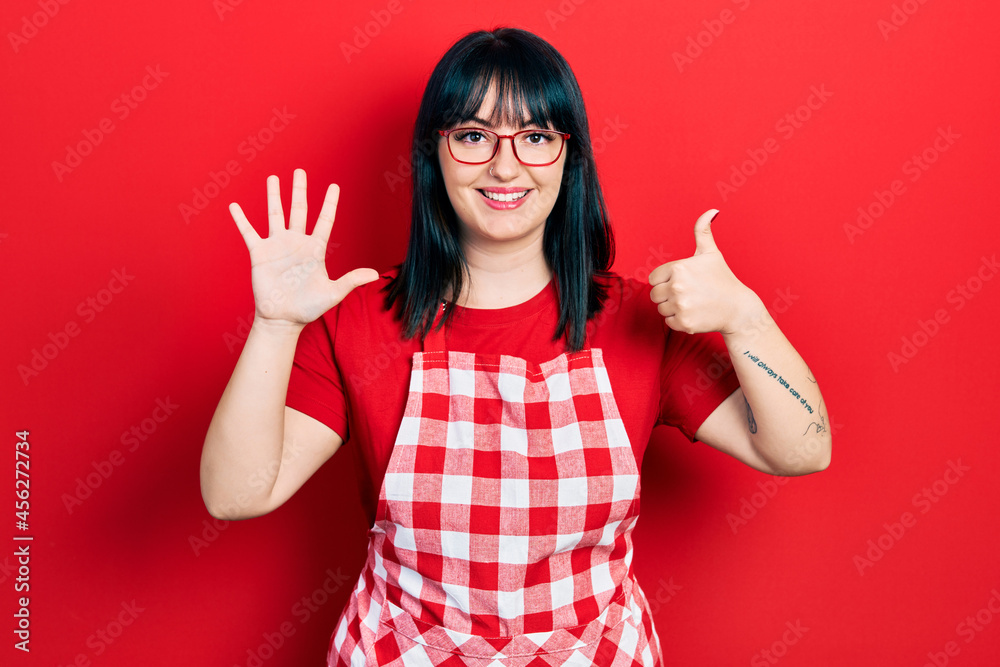Young hispanic woman wearing cook apron and glasses showing and pointing up with fingers number six while smiling confident and happy. Young hispanic woman wearing cook apron and glasses showing and pointing up with fingers number six while smiling confident and happy.
