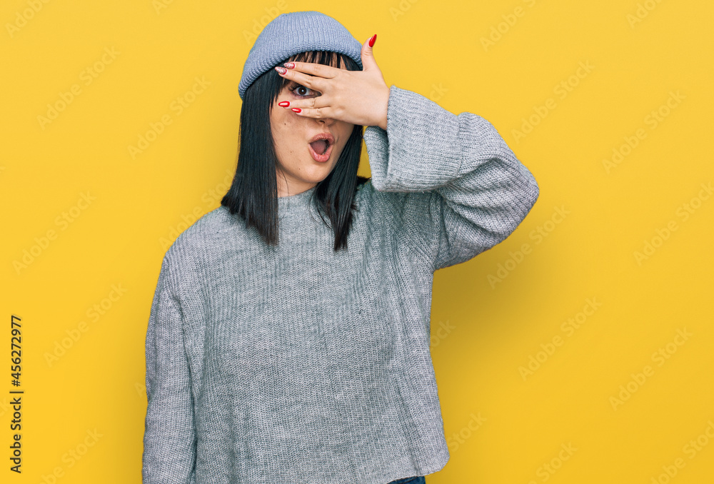 Young hispanic woman wearing cute wool cap peeking in shock covering face and eyes with hand, looking through fingers with embarrassed expression.