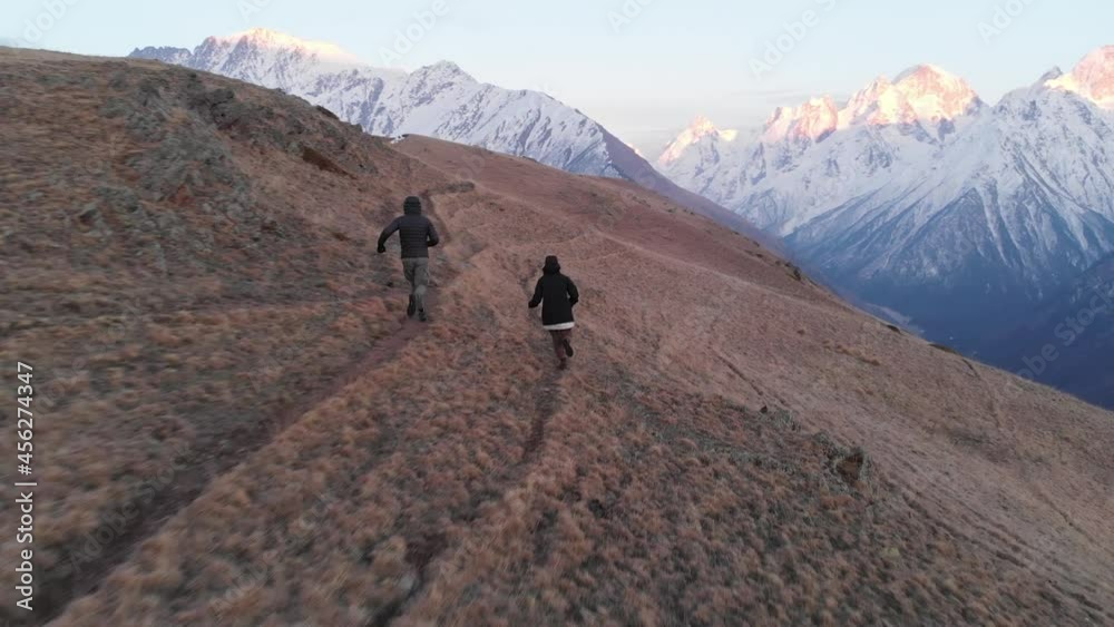A loving couple of climbers runs along a path on a mountain slope in the late evening against the backdrop of snow-capped mountains. A loving couple of climbers runs along a path on a mountain slope in the late evening against the backdrop of snow-capped mountains.