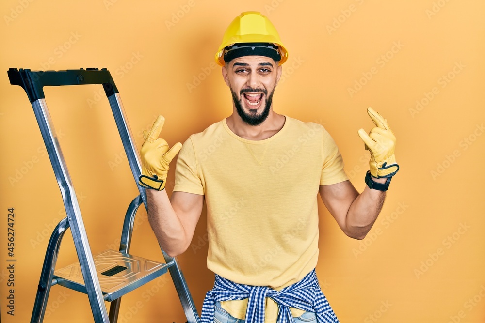 Handsome man with beard by construction stairs wearing hardhat shouting with crazy expression doing rock symbol with hands up. music star. heavy music concept. Handsome man with beard by construction stairs wearing hardhat shouting with crazy expression doing rock symbol with hands up. music star. heavy music concept.