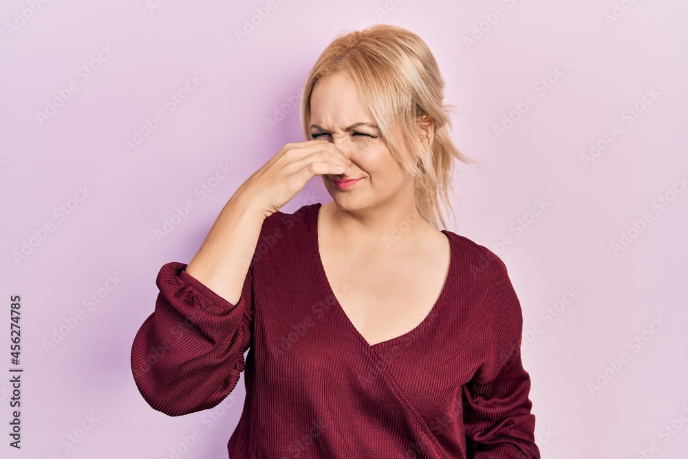 Young blonde woman wearing casual winter sweater smelling something stinky and disgusting, intolerable smell, holding breath with fingers on nose. bad smell Young blonde woman wearing casual winter sweater smelling something stinky and disgusting, intolerable smell, holding breath with fingers on nose. bad smell