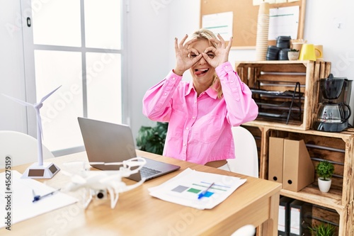 Young caucasian woman working at the office doing ok gesture like binoculars sticking tongue out, eyes looking through fingers. crazy expression.