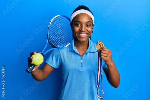 African american woman with braided hair playing tennis holding racket and ball and winner medal smiling with a happy and cool smile on face. showing teeth. © Krakenimages.com
