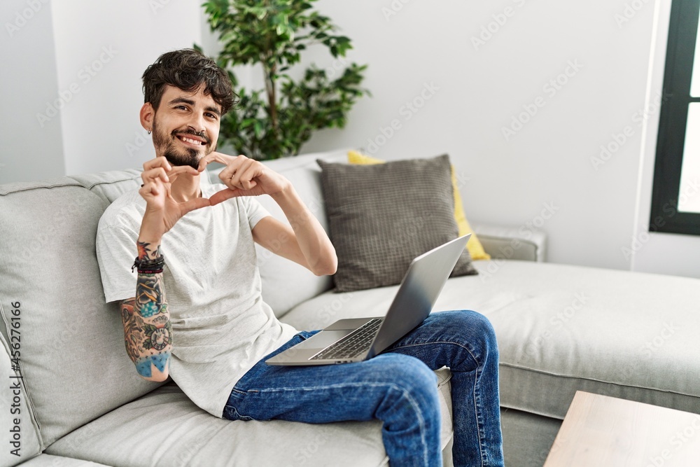 Hispanic man with beard sitting on the sofa smiling in love doing heart symbol shape with hands. romantic concept.