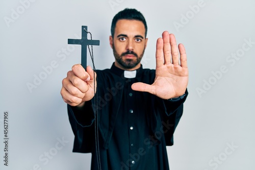 Fotografia Handsome hispanic priest man with beard holding catholic cross with open hand do