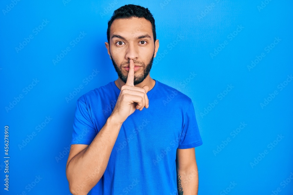 Hispanic man with beard wearing casual blue t shirt asking to be quiet with finger on lips. silence and secret concept.