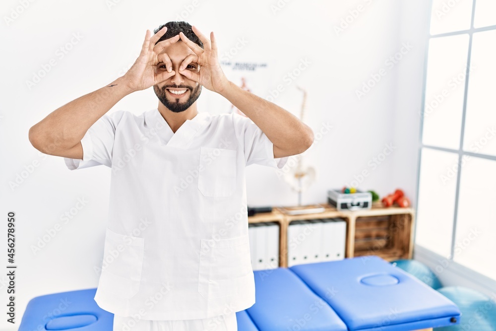 Young handsome man with beard working at pain recovery clinic doing ok gesture like binoculars sticking tongue out, eyes looking through fingers. crazy expression. Young handsome man with beard working at pain recovery clinic doing ok gesture like binoculars sticking tongue out, eyes looking through fingers. crazy expression.