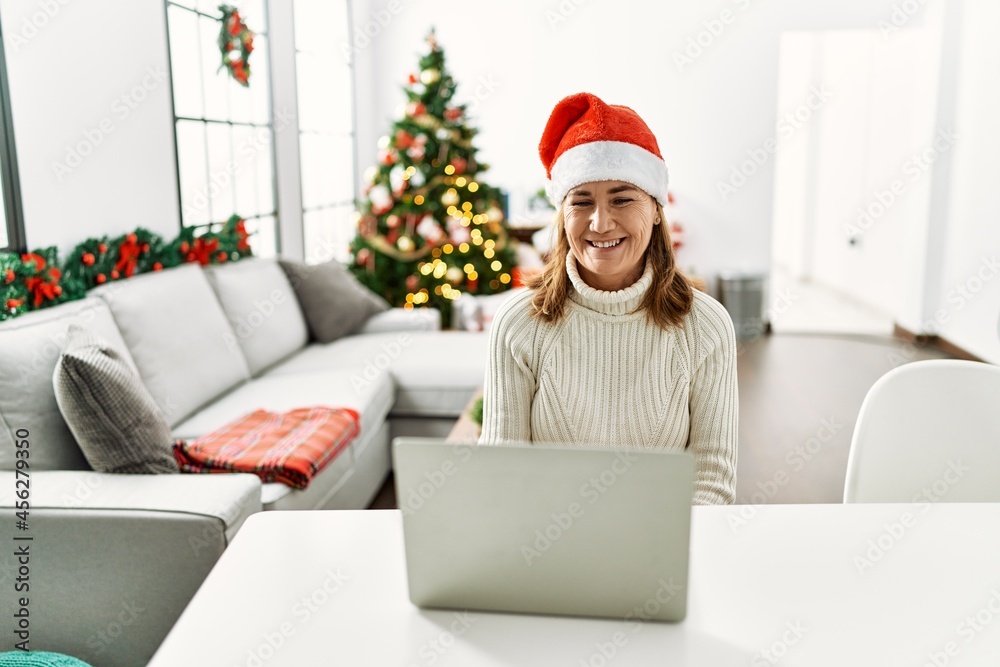 Middle age woman wearing santa claus hat using laptop looking positive and happy standing and smiling with a confident smile showing teeth Middle age woman wearing santa claus hat using laptop looking positive and happy standing and smiling with a confident smile showing teeth